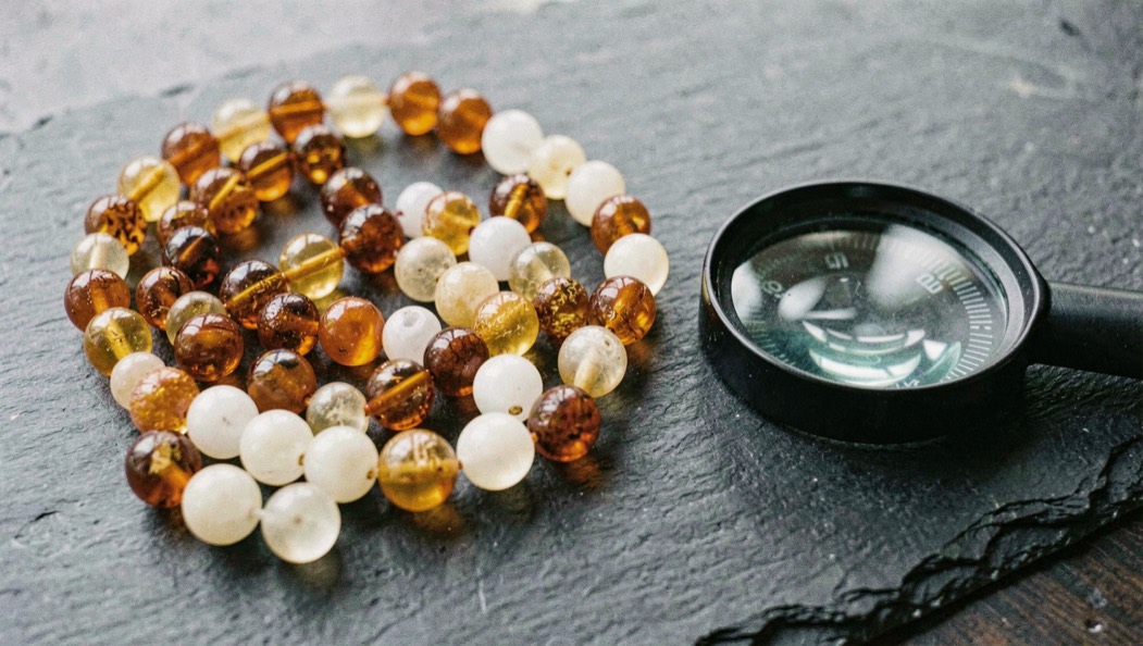 Assortment of amber beads in honey, cognac and milky white tones spread on dark slate beside a ten power loupe